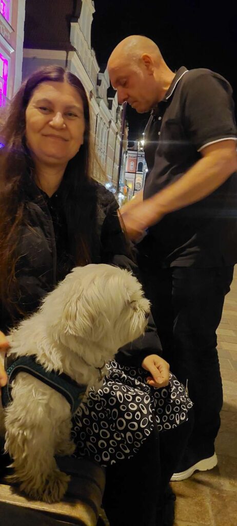 A dog is sitting with a woman and a man stands in the background in Poznan main square late at night